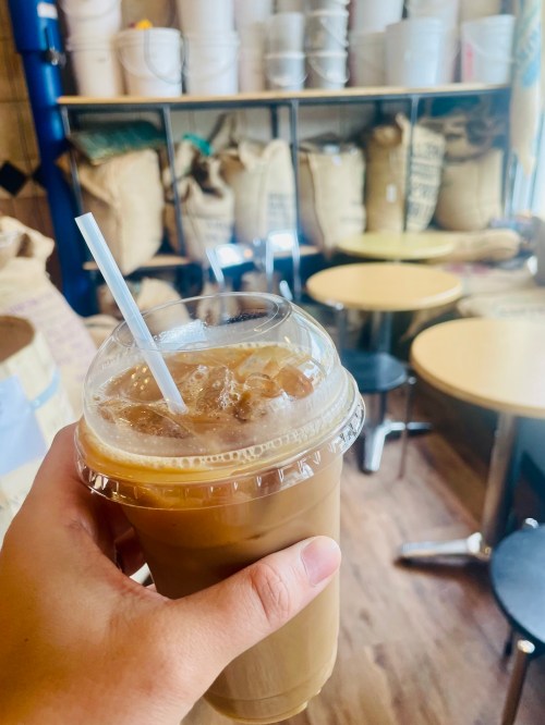A hand holding a plastic cup of iced coffee with a straw, set in a cozy café with wooden tables and bags in the background.