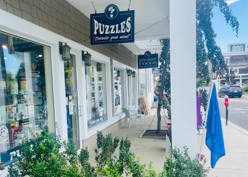 A storefront with a sign reading "PUZZLES" and a nearby sign for "LEWIS GOURMET FOODS," surrounded by greenery.