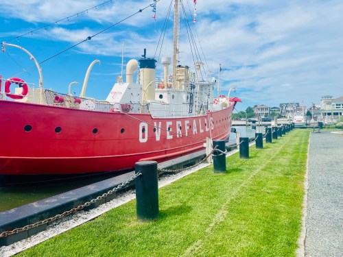 A red ship named "Overfalls" docked along a grassy waterfront with houses in the background under a blue sky.