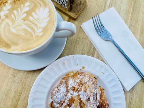 A cup of latte art coffee beside a pastry on a white plate, with a fork and napkin on a wooden table.