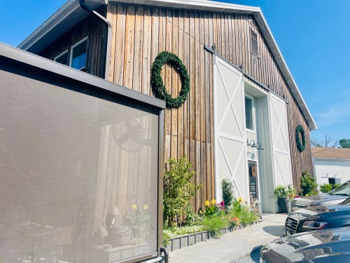 A rustic wooden barn with white doors, adorned with wreaths, surrounded by flowers and parked cars in the foreground.