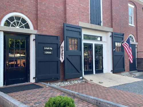 A brick building with large black doors, an "OPEN" sign, and an American flag displayed outside.