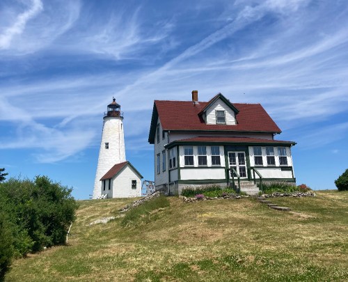 A white lighthouse beside a green and white house on a grassy hill under a blue sky with wispy clouds.