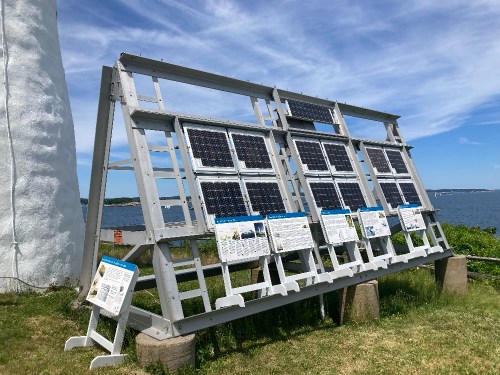 A solar panel display with informational signs, set against a backdrop of water and blue sky.