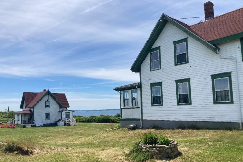 Two white houses with green trim sit on a grassy area near the water, under a clear blue sky.
