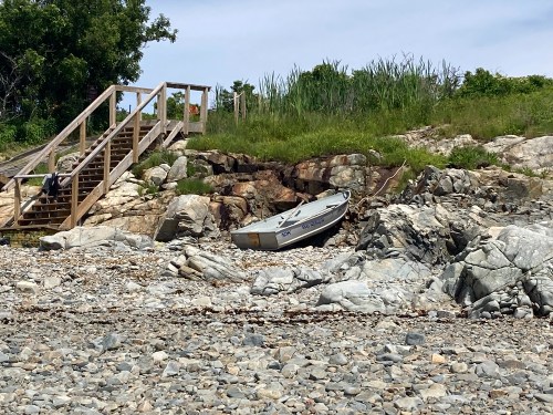 A small boat rests on rocky shore near wooden stairs, surrounded by grass and trees under a clear sky.