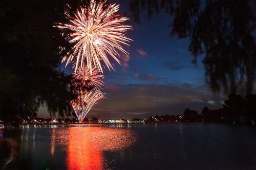 Colorful fireworks burst over a calm lake at night, reflecting vibrant lights on the water's surface.