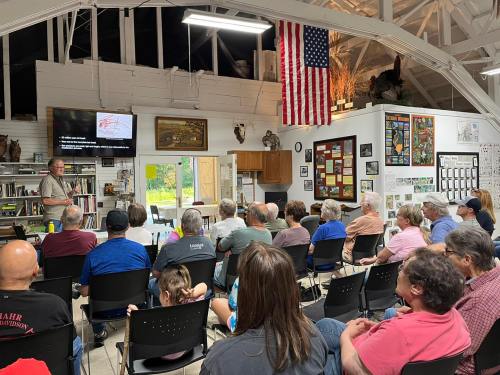 A group of people seated in a room, listening to a speaker in front of a screen, with an American flag displayed.