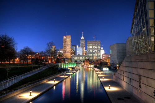 City skyline at dusk, reflecting in a calm waterway, with illuminated buildings and trees lining the banks.
