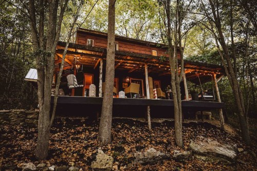 A rustic wooden cabin surrounded by trees, featuring a porch with chairs and warm lighting in a serene forest setting.