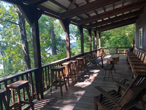 A wooden porch with several stools and chairs, surrounded by trees and a view of nature.