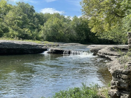 A serene river scene with a small waterfall, surrounded by lush greenery and rocky banks under a clear blue sky.
