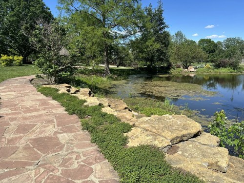 A stone path leads to a serene pond surrounded by greenery and trees under a clear blue sky.