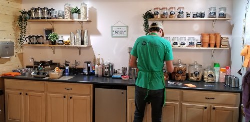 A barista in a green shirt prepares drinks at a cozy coffee shop counter with shelves of jars and utensils.