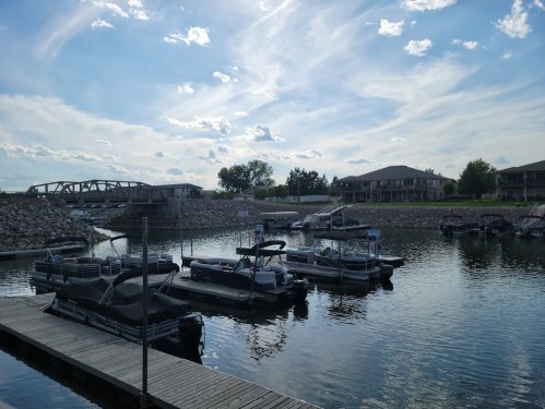 A serene marina with several boats docked, surrounded by calm water and a blue sky with scattered clouds.