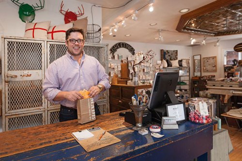 A smiling man stands behind a rustic counter in a charming gift shop, preparing a package for a customer.