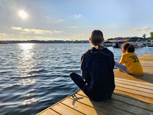 Two children sit on a wooden dock by a lake, watching the sun set over the water.
