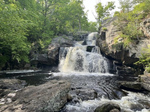 A serene waterfall cascades over rocky ledges, surrounded by lush green trees and a calm pool below.