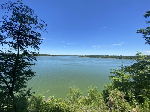 A serene view of a calm lake surrounded by lush greenery and a clear blue sky.