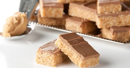 A close-up of two chocolate-covered rice crispy treats, with a stack of more treats and a spoonful of peanut butter in the background.