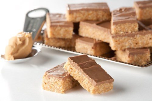 A stack of chocolate-covered rice crispy treats with a spoonful of peanut butter in the foreground.