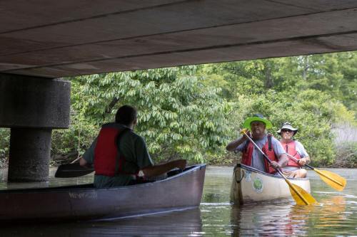 Two people in canoes paddle under a bridge, surrounded by lush greenery. One wears a wide-brimmed hat.