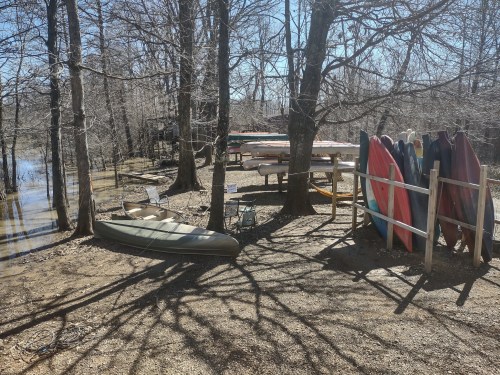 A serene outdoor scene with kayaks, surfboards, and a picnic table near a calm waterway surrounded by trees.