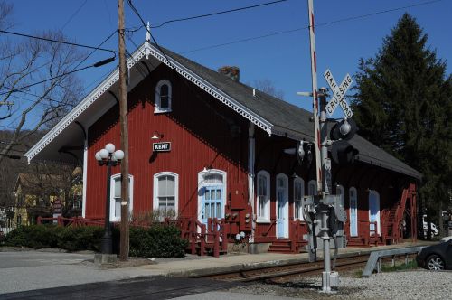 A red train station building with white trim, surrounded by trees and a railroad crossing sign in the foreground.