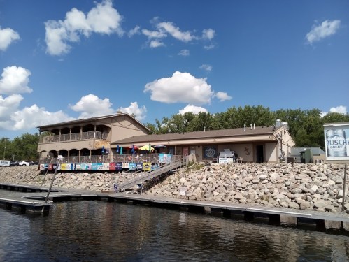 A waterfront building with a deck, surrounded by trees and clouds, featuring colorful flags and signage.