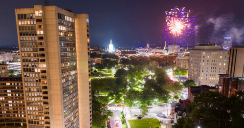 City skyline at night with fireworks above, showcasing parks and buildings illuminated in vibrant colors.