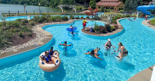 A water park scene with people floating on tubes in a winding pool, surrounded by greenery and a water slide in the background.