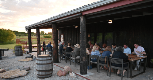 A group of people dining outdoors at a rustic venue during sunset, surrounded by nature.