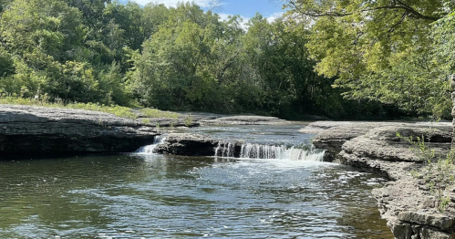 A serene river scene with a small waterfall, surrounded by lush green trees and rocky banks under a clear blue sky.