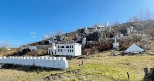 A hillside with miniature white buildings and structures against a clear blue sky, surrounded by dry grass and sparse trees.