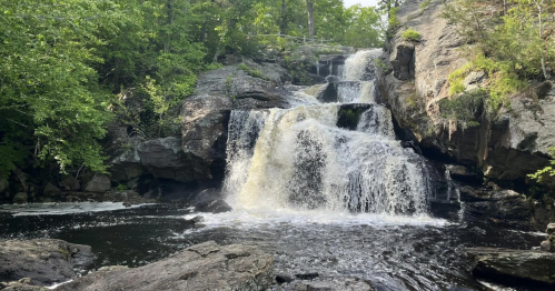 A serene waterfall cascades over rocky ledges, surrounded by lush green trees and a calm dark pool below.