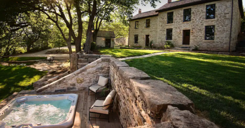 A serene outdoor scene featuring a hot tub, stone wall, and a historic house surrounded by trees and lush greenery.