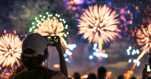 A person in a cap captures colorful fireworks on their phone, with a crowd watching in the background.