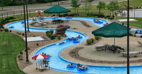 A winding lazy river pool with people floating on tubes, surrounded by green grass and shaded areas.