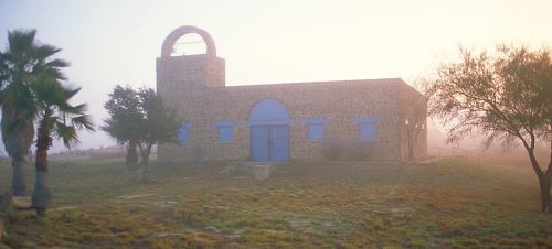 A stone building with blue accents surrounded by fog, featuring palm trees and a bell tower in the background.