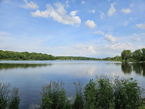 A serene lake surrounded by greenery under a blue sky with fluffy clouds reflecting on the water's surface.