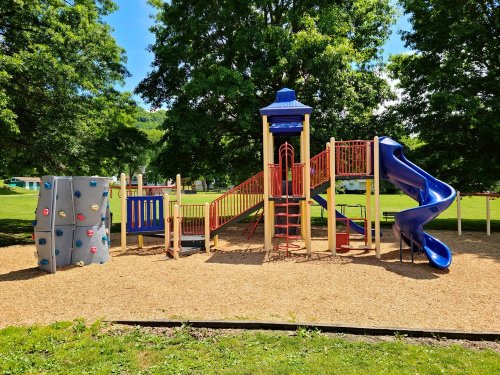 Colorful playground with a slide, climbing wall, and various play structures, surrounded by green trees and grass.