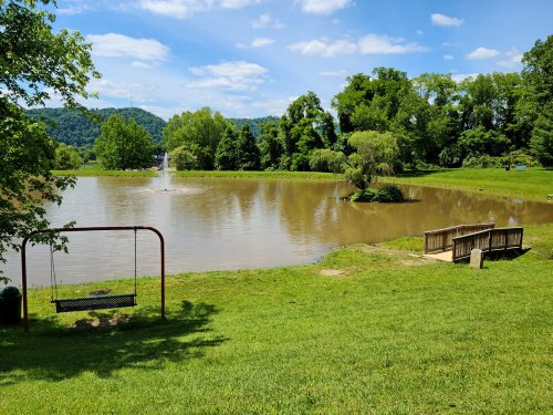 A serene park scene featuring a pond, a fountain, green grass, a swing set, and benches under a blue sky.