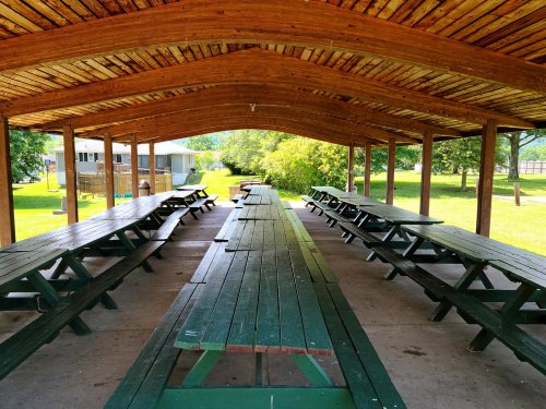 A spacious pavilion with wooden picnic tables under a sloped roof, surrounded by greenery and a pathway.
