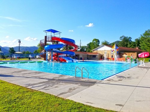 A sunny outdoor pool area featuring a large water slide, lounge chairs, and a grassy lawn.