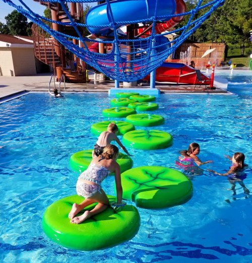 Children play on large green lily pad floats in a pool, with a water slide and climbing structure in the background.