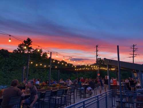 A lively outdoor dining area at sunset, with string lights and people enjoying their meals against a colorful sky.