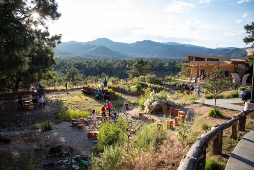 A scenic outdoor area with people enjoying nature, surrounded by mountains and greenery, featuring seating and gardens.