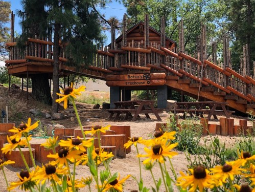 A wooden treehouse with a slide and picnic tables, surrounded by yellow flowers and trees.