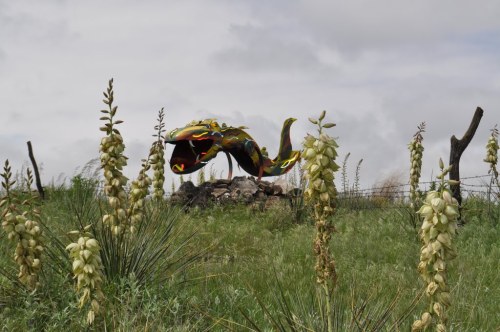 A colorful fish sculpture surrounded by tall green grass and flowering plants under a cloudy sky.