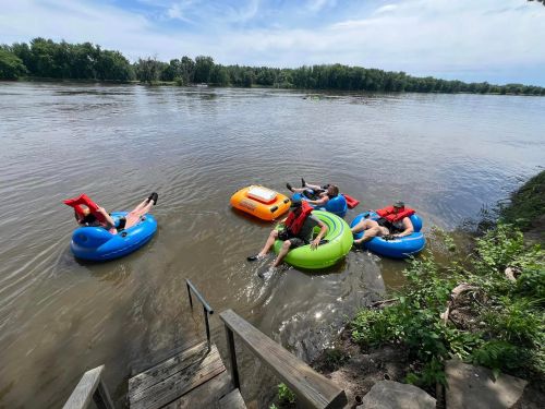 Four people relax on colorful inner tubes in a river, surrounded by greenery and a clear blue sky.